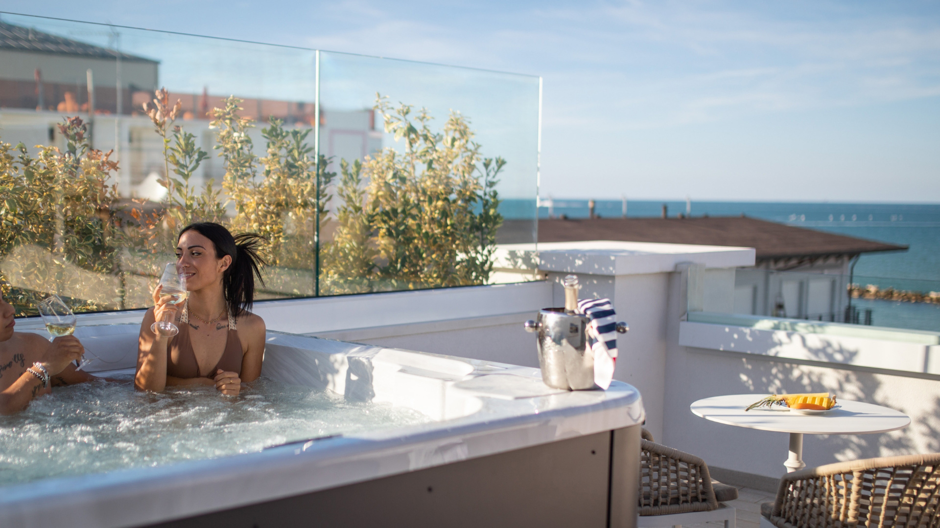 Due persone in jacuzzi con vista mare, brindano con bicchieri di vino bianco.