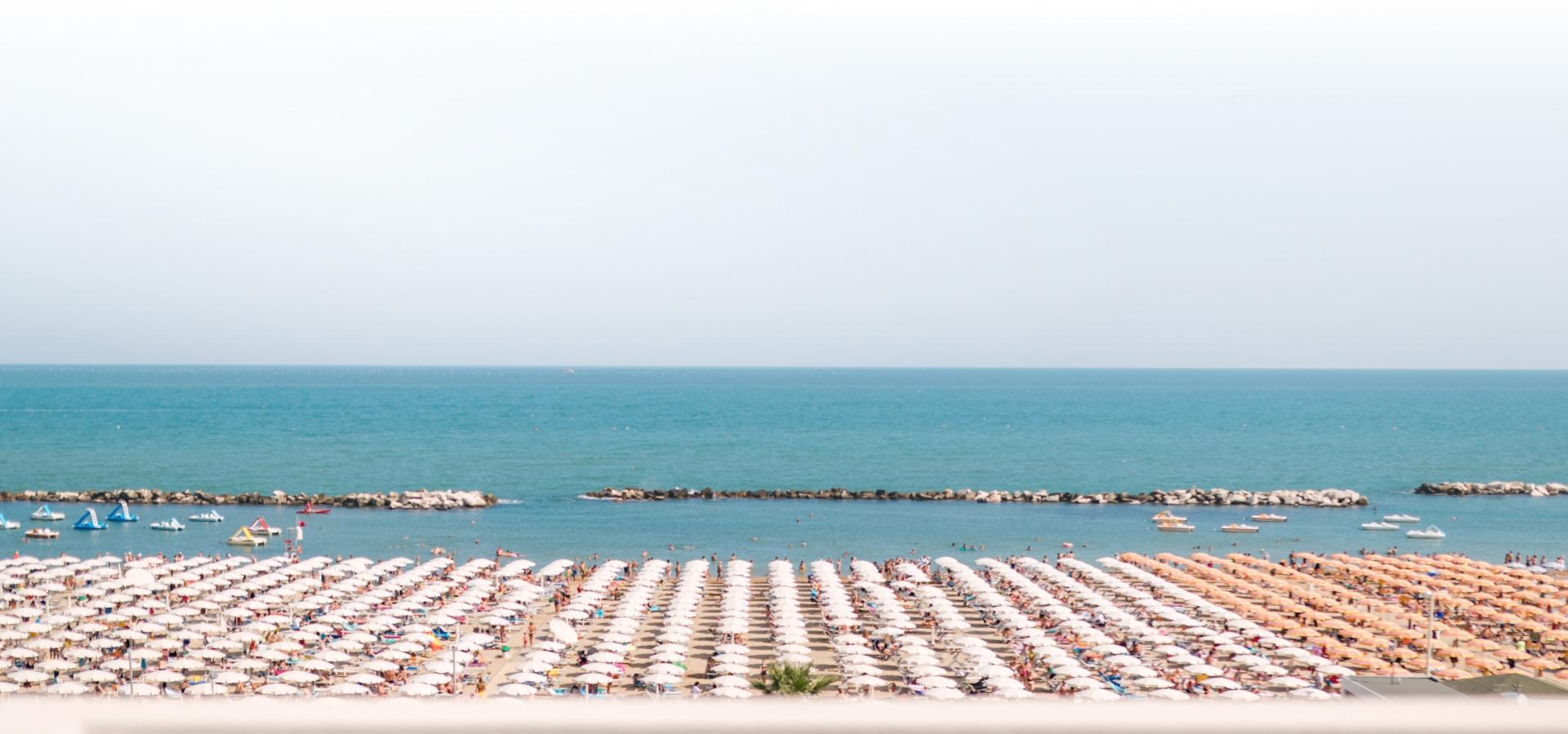 Spiaggia affollata con file di ombrelloni bianchi e mare calmo sullo sfondo.