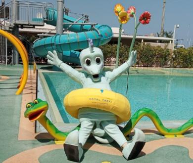 Mascotte sorridente con ciambella gialla e fiori, vicino a una piscina.