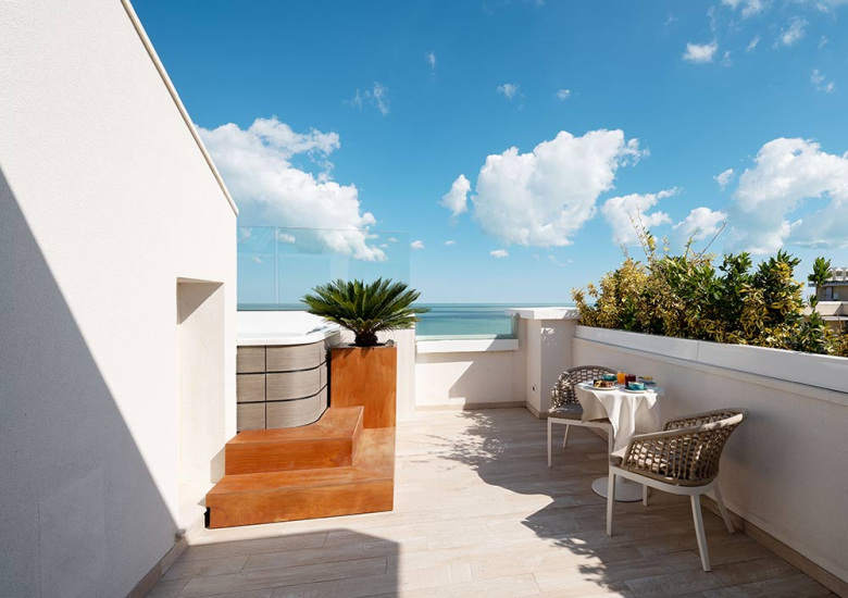 Terrazza con vista mare, tavolino con sedie, pianta e cielo sereno.