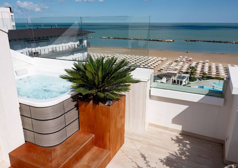 Terrazza con jacuzzi e vista mare, spiaggia con ombrelloni e cielo sereno.