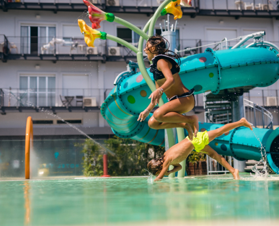 Bambini che saltano in piscina vicino a uno scivolo acquatico colorato.